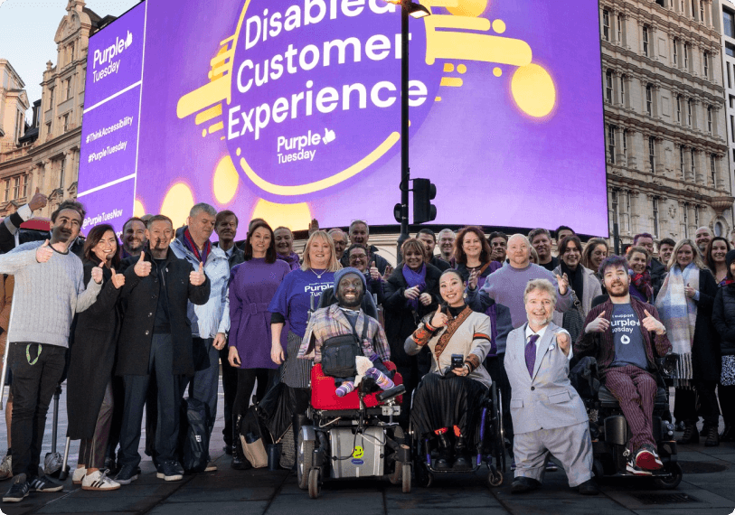 A diverse group of people, including individuals with visible disabilities and mobility aids, smile and pose together in front of a large digital billboard that reads "Disabled Customer Experience – Purple Tuesday." The group is gathered in a city square, promoting accessibility and inclusion.