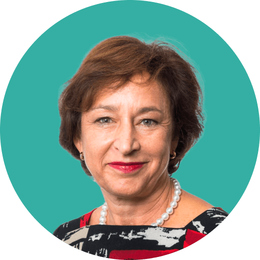 Professional headshot of a woman with short brown hair, wearing a patterned top, a pearl necklace, and red lipstick. She is smiling slightly and posed in front of a teal background.